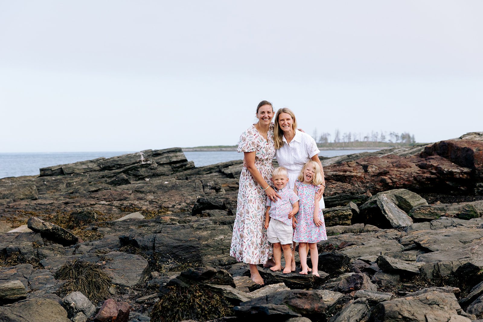 Two moms with young children smiling on rocky Maine coastline during family photo session