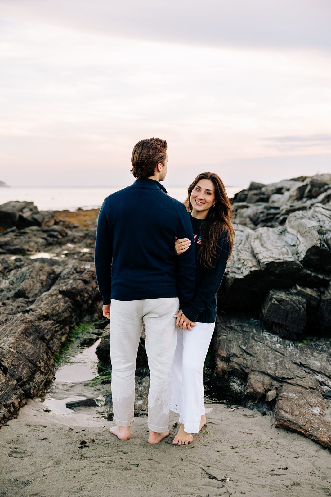 Engaged couple walking on rocky beach at Kettle Cove in Cape Elizabeth Maine during coastal engagement session